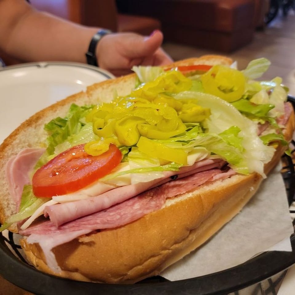 Large deli sandwich layered with ham, salami, cheese, lettuce, tomato, and banana peppers in a restaurant basket.